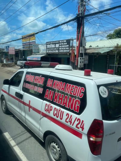 The image shows the side of a white ambulance with red lettering and a red stripe down its side. The ambulance has a red cross emblem on its door. The text on the ambulance, written in Vietnamese, indicates it is a "PHÒNG KHÁM ĐA KHOA MEDIC AN KHANG" which translates to "An Khang PolyClinic Medic". Below this, there is a phone number: "DT: 02762202969 0914 222227". Further down the side, in larger red letters, it says "CẤP CỨU 24/24", meaning "Emergency 24/24". The ambulance has two red flashing lights on its roof.
In the background, there are buildings with metal roofs and various signs, one of which reads "TRƯỜNG AN" and lists services such as "MÁY VI TÍNH MÁY IN, SCAN", "MÁY PHOTOCOPY", and "CAMERA QUAN SÁT", suggesting it might be an electronics or computer shop. There is also a Vietnamese flag visible. The scene appears to be on a street in Phường An Tịnh, Vietnam, with power lines overhead and trees in the distance. The sky is blue with scattered clouds. The ambulance is on a paved road with a white painted line.