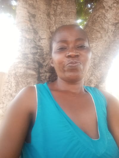 The scene, set in the city of Gudige Sabo, Nigeria, features a woman captured in a close-up, upper-body portrait. She has a dark complexion, with her dark hair pulled back. She is wearing a bright teal-colored sleeveless top with a deep V-neckline outlined by a thin white trim. Her lips are puckered into a distinct expression as she looks directly into the camera. The background is dominated by a large, light-barked tree trunk with visible texture and vertical furrows, suggesting a mature specimen that bifurcates above. Intense natural sunlight is evident on the right side of the frame, illuminating the environment, which appears to be outdoors. No other individuals or specific interactions are depicted.