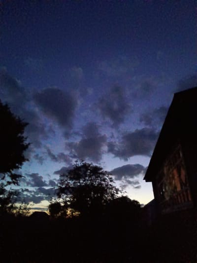 The image depicts an evening scene in Verkhnii Dvor, Russia. The sky is a mix of dark blue and gray, with numerous clouds. Silhouettes of trees and a building are visible in the foreground. The building is on the right side of the frame. The details of the building and trees are obscured due to the low light conditions. No people are visible.