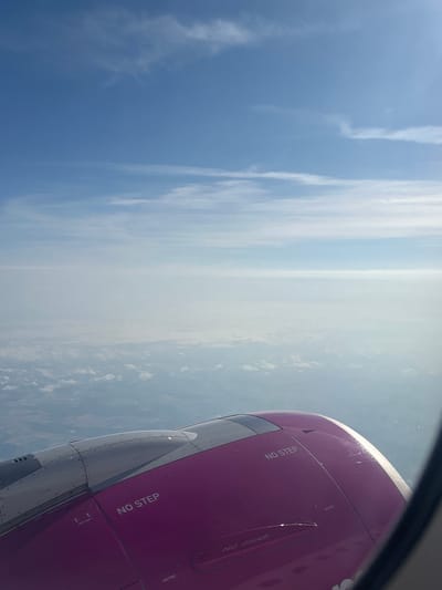 The image is an aerial view from an airplane window, looking downwards and outwards at the sky and landscape below. The foreground shows a portion of the airplane's engine cowling, painted in a vibrant magenta color, with "NO STEP" and "NO GRAB" stenciled on it. Above the engine, the gray metallic part of the wing is visible. The middle ground is filled with a layer of hazy clouds, obscuring the ground beneath. The background features a clear blue sky with wispy white clouds. The scene is a typical view from an airplane during flight. There is no discernible indication of the city of Echterdingen, Germany, in this image.