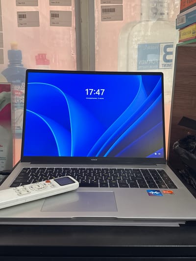 The image shows a laptop with a blue wallpaper and the time "17:47" displayed. A white remote control is placed on the laptop's touchpad. In the background, there are shelves with various items, including what appears to be bottles and boxes. The overall setting suggests an indoor environment, possibly a store or a room with merchandise. There is no indication of the location being Yovon, Tajikistan, or any other specific city.