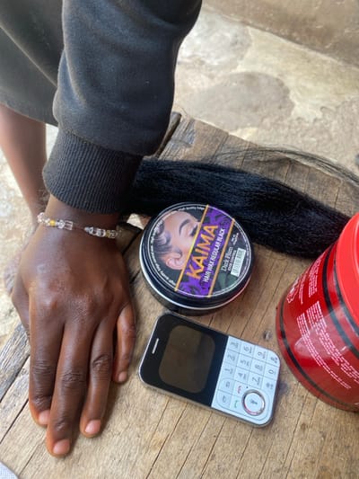 The scene is set on a light-brown wooden surface in Idiroko, Nigeria, under bright outdoor illumination. A human left arm, clad in a dark, ribbed-cuff long-sleeved garment, rests partially visible. A clear and yellow beaded bracelet adorns the wrist. Arranged on the wooden surface are several items: a bundle of black synthetic hair, a black circular tin of "KAIMA HAIR WAX REGULAR BLACK - Dark Plum" for edge control, a large red cylindrical plastic jar with a partially visible label indicating "STYLING GEL," and a silver-colored feature phone with a numerical keypad, small screen, and a dedicated flashlight button. The phone's screen is dark. No explicit actions or interactions are depicted beyond the static arrangement of objects.