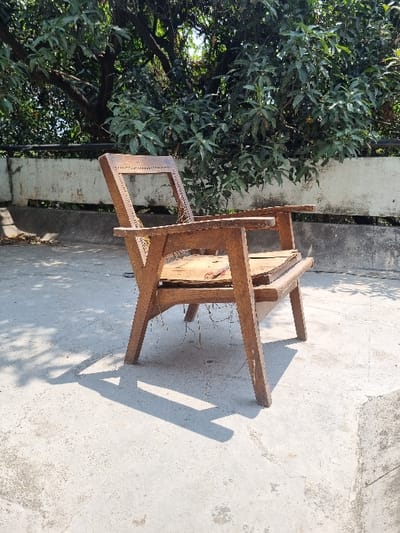 A wooden chair sits on a concrete surface, with a white wall and a lush green tree behind it. The chair's seat is made of woven material and appears to be missing a cushion. The location is in Lucknow, India at coordinates 26.915, 80.944. The chair is facing away from the viewer. The chair is positioned under a tree, casting a shadow on the concrete surface. The tree's leaves are green and lush, creating a serene environment.  The chair appears to be slightly weathered, possibly due to exposure to the elements.  There are no people or objects in the immediate vicinity of the chair.  The surrounding environment appears to be residential or semi-rural.
