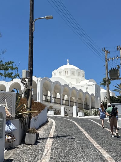 This is a vertical, eye-level, outdoor shot of a white domed church in Thira, Greece, under a clear blue sky. The church is the primary subject, featuring a prominent dome topped with a cross and numerous arched porticos along its facade. A cobblestone street, marked with white lines, leads towards the church.

In the foreground, on the left side, a white wall displays several patterned bags hanging on a rack. Next to it, a stone planter holds a spiky plant. A dark wooden utility pole with electrical equipment and a street lamp stands on the left side of the street, with power lines extending across the sky.

On the right side of the street, another utility pole supports a large transformer and more power lines. Three individuals are walking away from the camera towards the church. The person in the front wears a blue dress and is partially obscured. The person in the middle wears a green shirt and tan shorts, carrying a black backpack. The person behind them wears a white hat and is also carrying a backpack.

The environment is sunny, indicated by the bright light and shadows. The architecture suggests a Mediterranean setting, consistent with Thira, Greece.