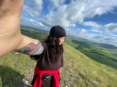 A person wearing a black cap, dark brown sweatshirt, and a red jacket tied around their waist is standing on a grassy hilltop, taking a selfie. The person has long, dark, wavy hair and a tattoo on their left arm. A fanny pack is visible around their waist. The background shows a vast, green landscape with rolling hills under a partly cloudy blue sky. The location is identified as Andreevka, Russia.