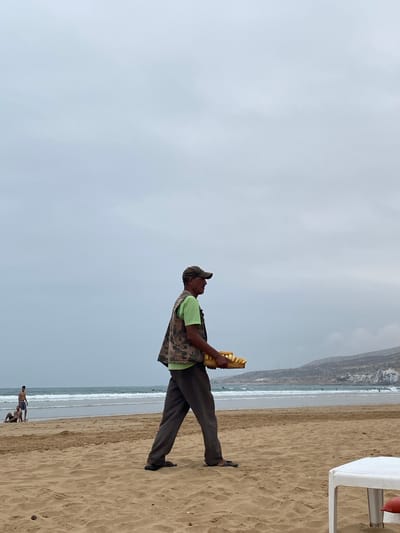 An eye-level, medium outdoor shot shows a man holding baguettes walking on the beach in Taghazout, Morocco. The man, possibly a street vendor, is facing right and holding a stack of at least 6 yellow baguettes. He is wearing a green t-shirt, camo vest, brown pants, a hat, and sandals. In the background is a sandy beach, the ocean with waves, and a hazy mountain-like structure with building on top. A white chair is visible on the right, in the foreground. In the distance are people swimming in the ocean. The sky is gray and overcast.