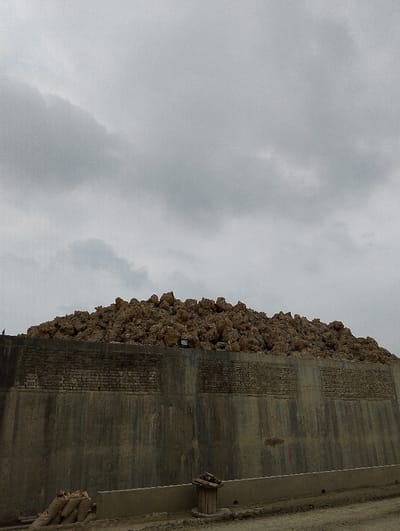 A large pile of rocks sits atop a concrete retaining wall in the city of Pangasia, Bangladesh. The wall has a brick pattern near the top. The scene is overcast with clouds and the ground is dirt. There are a few bags and a wooden trash can in the foreground.  The coordinates 22.694, 90.601 place the location in the area surrounding Dhaka, Bangladesh.