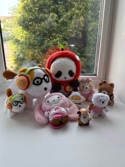 A collection of stuffed toys is arranged on a white surface near a window. The toys include a panda in a red hood, a white dog wearing a burger-themed hat and glasses, a pink Hello Kitty plush, a small yellow chick, a teddy bear with a pink bow, a brown teddy bear, a white dog holding a baseball bat, and a small gnome figure. The window shows an exterior view of green trees and foliage. The image appears to have been taken indoors with natural light. There is no verifiable location in Severnyi, Russia.