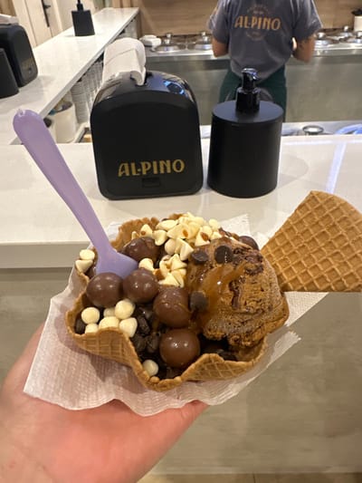 An ice cream cone with a waffle cookie garnish is displayed. The cone holds chocolate ice cream, caramel drizzle, chocolate chips, white chocolate chips, and chocolate-covered malt balls. A purple spoon is inside the cone. The cone sits on a white napkin held by a hand. A machine with the "Alpino" logo is nearby, as is a dispenser. An employee with an "Alpino" shirt stands next to the machine.