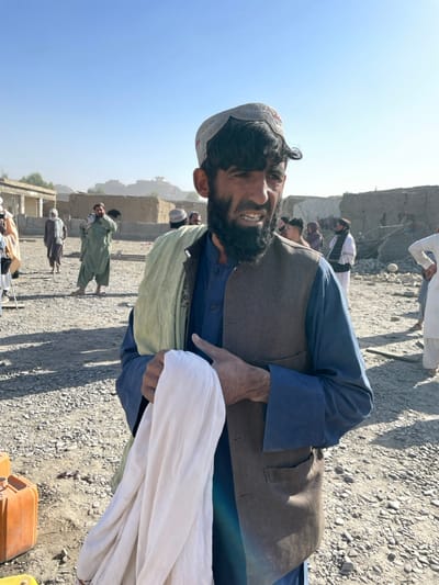 This image captures an outdoor scene in Spin Boldak, Afghanistan, under a clear, bright blue sky on a sunny day. The primary subject is a male, mid-adult, positioned in the foreground, facing slightly to his right. He has dark hair, a full dark beard, and a mustache, and wears a light-colored, possibly crocheted, skullcap. His attire consists of a dark blue traditional tunic and trousers, a dark brown vest, and a light green scarf draped over his left shoulder. He holds a folded white piece of fabric in his hands at chest height. His mouth is slightly open, revealing his teeth.
The ground is dry, dusty, and covered with small stones or gravel. In the lower-left corner, an orange jerrycan is partially visible. The background contains multiple other males, dressed in traditional attire, standing or walking on similar terrain. To the left, a light-colored building with distinct columns appears partially intact. To the right, various structures are visible, some appearing dilapidated, damaged, or partially collapsed, constructed from mud-brick or concrete. Distant, low-lying structures or hills are discernible on the horizon.