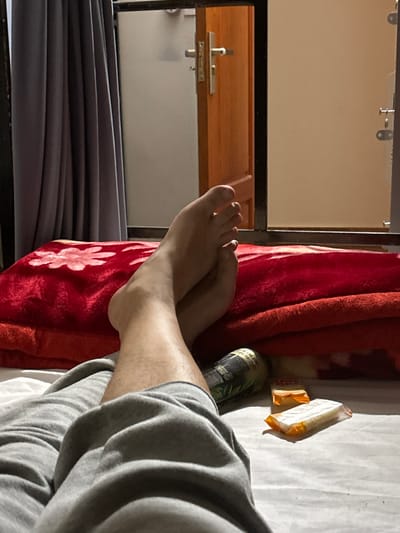 A first-person view captures the lower legs and bare feet of an individual, clad in grey sweatpants, resting on a bed. The feet are crossed at the ankles. The bed features a white sheet covered by a red blanket with a subtle lighter-toned pattern, possibly floral. An orange item, likely a pillow or folded blanket, is visible beneath the red blanket. Adjacent to the person's legs on the bed are a dark green plastic bottle or cup and two individually wrapped, rectangular items in orange and white packaging. In the background, a wooden door with a silver handle is slightly ajar, revealing a light beige wall or door beyond it. To the left of the wooden door, grey curtains are partially drawn. The scene is an indoor setting, likely a hotel or guesthouse room in Phong Nha Commune, Vietnam.