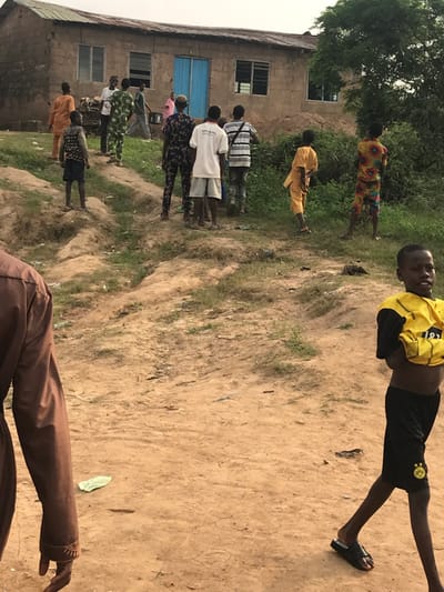 People stand in a dirt yard outside a building with a blue door and several windows. The group comprises mostly youths dressed in varied casual clothing, with some wearing patterned shirts and shorts. A young man in a yellow and black soccer jersey and sandals is prominent in the foreground. The ground is bare and uneven, with a few scattered leaves. A partial view of a person in a brown jacket is at the lower edge of the frame. Trees can be seen in the background near the building.