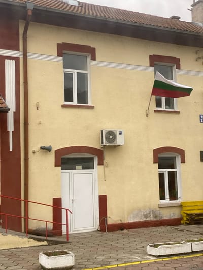 This image depicts the exterior of a two-story building in the city of Zemen, Bulgaria, under an overcast sky. The building features a textured, light yellow facade with dark reddish-brown trim around windows, doors, and along the base and roofline. The roof is covered with reddish-brown tiles.
On the upper floor, two rectangular windows with white frames are visible. Between them, mounted to the wall, is a pole displaying a Bulgarian flag, which unfurls to the right, showing horizontal bands of white, green, and red. A third, smaller window with a white frame is situated below the flag, on the ground floor.
Between the main windows on the ground floor, a white modern door with a semicircular arch is visible, framed by the same reddish-brown trim. Above this door, an external air conditioning unit is mounted on the yellow wall. To the left of the door, a gray speaker projects from the wall. A reddish-brown downspout runs vertically along the left edge of the building facade.
The ground area in front of the building is paved with interlocking gray blocks, outlined by a yellow painted line. A red metal handrail descends towards the left from the building. Two square, white concrete planter boxes containing sparse vegetation are positioned on the paved surface. To the far right, a yellow wooden bench is visible, resting on a stack of pallets or similar objects. No individuals are present in the scene. The building shows some signs of wear and discoloration near the base.