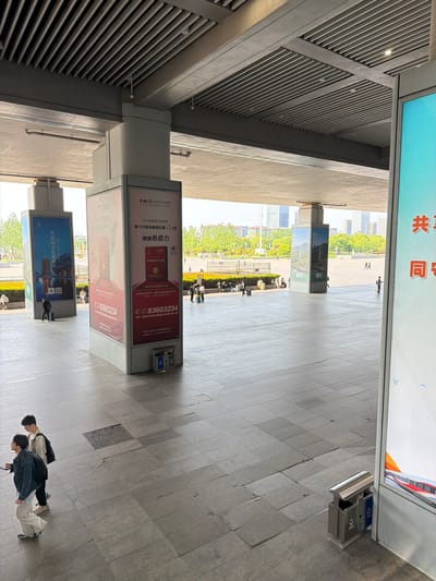 This image captures a high-angle view of a modern, covered public walkway or underpass, leading out to an open plaza or park area in Yuhuatai District, China. The scene is illuminated by bright daylight, suggesting clear weather.
In the foreground, two individuals are seen from above, walking on the grey paved floor. Further along the corridor, several other people are visible, some standing near structural pillars, others seated on benches in the outdoor area. The architecture features a distinctive grey slatted ceiling and robust pillars that support large, illuminated billboards.
The billboards display various advertisements and information. One prominent blue billboard features the text "牛首山文化旅游区" (Niushoushan Cultural Tourism Zone). Adjacent red billboards advertise "中科牌番茄红素胶囊" (Zhongke brand Lycopene Capsules) with details such as "中科SINCE1984," "每100克含番茄红素≥7.0克" (≥7.0 grams of lycopene per 100 grams), and "增强免疫力" (Enhance immunity). Contact information, including the phone number "025-83603234" and an address in Nanjing, is also visible. A large overhead sign in the background displays the character "共同" (common/together).
Beyond the covered area, lush green trees and distant buildings indicate an urban outdoor environment. Two dual-compartment trash bins are placed near the billboards. The overall setting appears to be a busy public thoroughfare, possibly connected to a transportation hub or a large commercial/cultural complex.