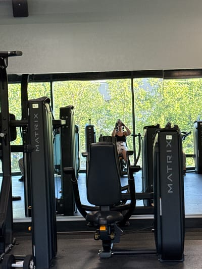 In a gym in the City of Yonkers, United States of America, a person is seen exercising. The person, wearing a black tank top and patterned shorts, is seated on a machine and holding a phone up, likely taking a selfie. Multiple pieces of gym equipment are visible, including a machine with the "MATRIX" logo prominently displayed. The gym features large windows with a view of lush green trees, suggesting an outdoor natural setting beyond the glass. The floor is a dark, gym-style surface, and the overall lighting indicates a daytime scene. The perspective is from within the gym, looking towards the window and the person exercising.