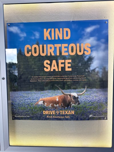 A poster promoting safe driving is displayed behind glass. The poster features a Texas Longhorn cow lying in a field of bluebonnets, with a calf partially visible behind it. The sky above is a bright blue with white clouds. Large orange text at the top of the poster reads "KIND COURTEOUS SAFE". Below the cow, smaller orange text reads "DRIVE like a TEXAN. Kind. Courteous. Safe." A website address "DriveLikeATexan.com" is visible in white text on the left side of the poster. The Texas Department of Transportation logo is present on the right side of the poster. The poster is framed and mounted on a wall. The reflection of trees and a street with a traffic signal is visible on the glass in front of the poster. The image was taken in Sonora, United States.