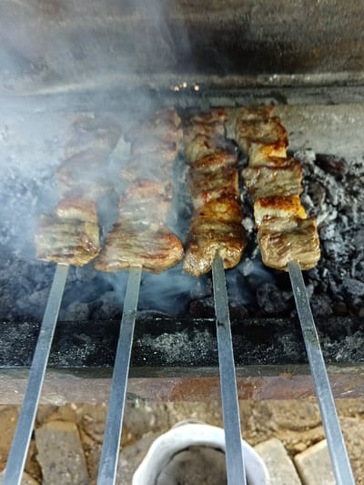 The image depicts multiple skewers of meat being grilled over hot coals in Kasri Arifan, Uzbekistan. Four metal skewers, each laden with approximately five to seven pieces of cubed or sliced meat, are positioned horizontally above a bed of glowing charcoal embers and ash. Wisps of white smoke rise from the cooking meat and coals. The meat pieces exhibit varying degrees of browning and charring. The grilling apparatus appears to be a dark, rectangular metal brazier or mangal. Below the brazier, the ground consists of dirt or rough concrete with visible brick fragments and a partial view of a small, white cylindrical container.
