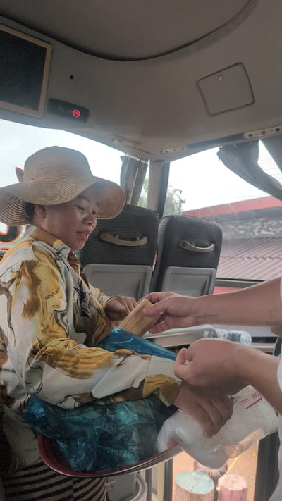 A woman wearing a wide-brimmed straw hat and a patterned shirt is seated inside a vehicle, possibly a bus, as indicated by the seating arrangement and interior features. Her hands are visible, and she appears to be holding a bundle of what looks like dried reeds or bamboo. Another person's hands are reaching towards her, offering or receiving a white plastic bag. The interior of the vehicle features dark gray seats with handles and a beige ceiling. A no-smoking sign with a red circle and a diagonal line is illuminated on the ceiling. The background shows windows with curtains and glimpses of the outside environment, suggesting an urban or semi-urban setting. The presence of wood stumps in the lower right corner of the image might suggest an outdoor or roadside location.