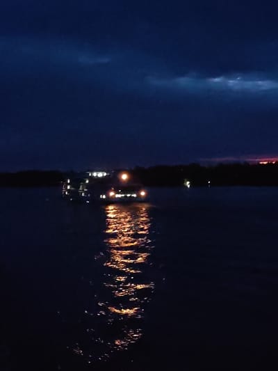 The image depicts a dark, night scene on the water, likely a lake or river. A multi-tiered boat, illuminated with multiple lights, is centrally positioned in the scene. The lights from the boat reflect and create a shimmering pathway on the water’s surface toward the viewer. The background shows a treeline of dark, silhouetted trees. Above, the sky is a gradient of dark blues and blacks with some faint patches of lighter shades and a glimpse of a fiery sunset on the horizon. It's unknown if this is Kadnikovo, Russia.