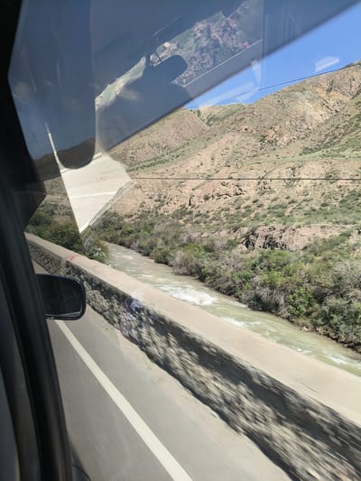 The image captures a scenic view of a river flowing through a mountainous landscape, likely in the vicinity of Cholok, Kyrgyzstan. The perspective suggests the photo was taken from a moving vehicle, possibly a car, looking out through the side window.
In the foreground, the edge of a car window and a side mirror are visible on the left. A concrete barrier runs along the side of a road, with a white line marking the edge of the asphalt. The road itself appears to be moving due to the motion blur.
Beyond the barrier, a river with churning, light-colored water flows from left to right. The banks of the river are lined with dense green vegetation, including trees and bushes.
In the background, rugged, barren mountains rise steeply. The slopes are covered in sparse vegetation and appear to be composed of dry soil and rocks. The sky above is a clear, bright blue. Reflections of the interior of the vehicle, including what might be seats or other passengers, are visible on the car window, adding a layer of depth and context to the scene. The overall impression is one of natural beauty and rugged terrain, characteristic of mountainous regions.