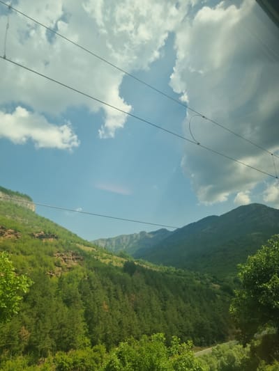 The image displays a scenic view of a mountainous landscape with lush green vegetation under a partly cloudy sky. In the foreground, the slopes are densely covered with trees, transitioning to rockier, less vegetated terrain higher up. Distant, hazy mountain ranges are visible in the background. Overhead, power lines are strung across the frame, and there is a reflection on a surface, possibly a window, in the upper right corner. The scene appears to be captured from a moving vehicle, given the presence of the power lines and the slight blur in some areas. The location is identified as Ochindol, Bulgaria.