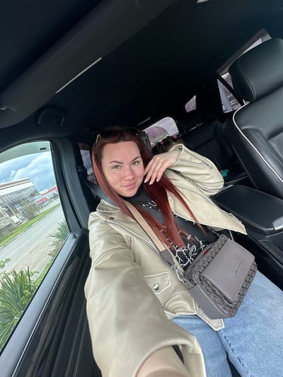 The image shows a woman with auburn hair lying in the passenger seat of a car. She is wearing a light-colored jacket and jeans, and a grey Michael Kors purse is positioned on her lap. The car is parked, and a view of Proletarskii, Russia can be seen through the window, showing buildings and greenery.