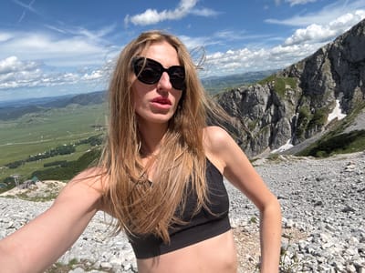 A woman with long blonde hair, wearing large black sunglasses and a black sports bra, is taking a selfie from a mountainous location. She is positioned in the foreground, with her right arm extended towards the camera. In the background, a vast, green valley with scattered houses and a ski lift stretches out, bordered by distant mountains. A steep, rocky mountainside with patches of snow and vegetation rises sharply on the right side of the frame. The sky is blue with scattered white clouds. The specific location is Pašina voda, Montenegro.