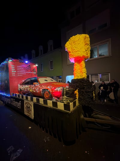 A carnival float is displayed at night on a street in Wasserbillig, Luxembourg. The float features a central orange-red car, stylized as Lightning McQueen from Disney's "Cars" and illuminated with white and red string lights. To the car's left, a red banner exhibits a schedule with dates and locations, including "WAASSERBËLLEG" and "CLIÄRRWER MAATSKAVALKAD," alongside the text "Landjugend Cliärref." To the right of the car, a large, yellow and red sculpture, appearing to be made of crumpled material and resembling a mushroom cloud, is mounted on the float. The float itself has a black base with white and black checkerboard trim and a dark pleated skirt. In the background, multi-story buildings with illuminated windows line the street. Several individuals, including an adult holding a white bag and a child, stand on the sidewalk to the right of the float. Small pieces of debris are scattered on the dark street pavement.