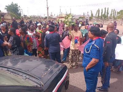 The scene is a protest taking place in Ungwan Tanko, Nigeria. A large group of people, mostly women, are gathered in front of a black car. There are several people holding signs, one of which has a picture of a man on it. In the foreground, two security officers are standing near the edge of the group. One officer is wearing a blue uniform with gold braid and the other officer is wearing a black uniform with the word "SECURITY" printed on it. Several people are interacting, some are speaking with each other, and others are holding signs. There are several trees in the background, and there is a factory behind the trees. The environment is a paved street, and there are trees and a factory behind the people. The protestors are standing in front of a black car.
