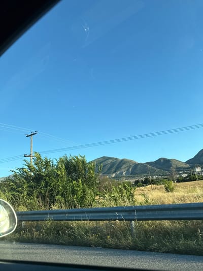 The image depicts a roadside view with a clear blue sky and rolling hills in the background. A metal guardrail runs horizontally across the lower portion of the frame, separating the road from a grassy embankment. Lush green bushes and dry, golden grass are visible on the embankment, with a wooden utility pole and power lines stretching across the scene. In the distance, several buildings are scattered among the hills, suggesting a semi-rural or suburban area. The overall impression is one of a sunny day in a natural landscape, possibly on the outskirts of a town. The presence of the utility pole and power lines indicates infrastructure typical of a developed area.