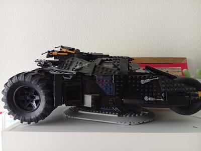 This media file displays a side profile of a black LEGO vehicle, resembling a Batmobile, placed on a white shelf. The vehicle is constructed from various black LEGO bricks, with a large, rugged black tire on the left side. The front of the vehicle is partially visible, showing orange and black LEGO pieces that appear to be weapons or decorative elements. The body of the vehicle is detailed with numerous small circular bumps, suggesting armor plating, and has some transparent blue LEGO bricks interspersed within the black structure, adding a touch of contrast. A grey, disc-like base with stud details supports the front portion of the vehicle. The background is a textured white wall. The lighting appears to be even and diffused, suggesting an indoor setting, possibly a room with artificial light or indirect natural light. There is no visible text within the image. The overall impression is of a meticulously built LEGO model on display.
