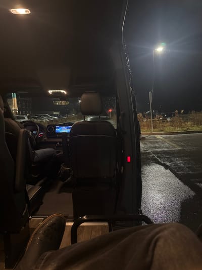 Captured from inside a car at night, the scene shows rain-slicked asphalt and a distant streetlight. The dashboard and steering wheel are visible, along with a glimpse of the infotainment system. A person's leg, clad in jeans and a dress shoe, is partially in frame on the right. Exterior buildings with lighted windows can be seen.