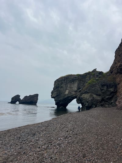 The image depicts a coastal landscape, likely a beach in Gvozdevo, Russia. The scene features a large rock formation with a natural archway. In the background, there is a smaller rock formation, also with an arch, visible in the water. A person stands near the base of the larger arch. The foreground is dominated by a rocky beach. The sky is overcast.