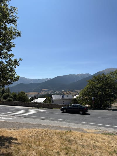 A black sedan is driving on a paved road next to a crosswalk. The road is lined with a stone wall and dry grass. In the background, there are houses and buildings, and behind them, a mountain range under a clear blue sky. The image was taken in La Cabanasse, France.