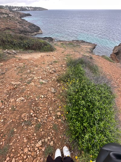 The image shows a cliffside view in Llucmajor, Spain. The ocean is visible to the left, with rocky cliffs and vegetation along the edges. The ground is primarily red dirt and small rocks, with patches of green vegetation including bushes and small yellow flowers. Part of a person's legs and white shoes are visible in the lower right corner of the image. A portion of a black object is visible above the shoes.