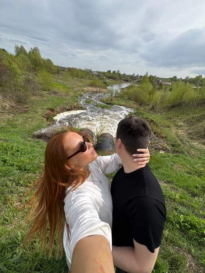 Two people are next to a small waterfall. A person in a black shirt has a hand on the neck of a person in a white shirt and sunglasses, and the person in the white shirt is looking up at the person in the black shirt. They are in Nozhovka, Russia.