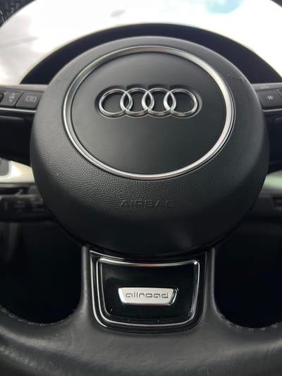 A close-up, eye-level shot focuses on the steering wheel of an Audi vehicle, likely parked in Sânnicoară, Romania. The emblem of four interlocking rings, the Audi logo, is prominently displayed in the center of the steering wheel. Below the emblem, the word "AIRBAG" is embossed. To the lower part of the steering wheel, a small rectangular badge with the text "allroad" is visible. The steering wheel itself is black and appears to be made of leather. On the left side of the steering wheel, some controls with symbols and the letters "OK" are partially visible. The background is indistinct, suggesting an indoor or shaded environment, possibly a garage or a parked car.