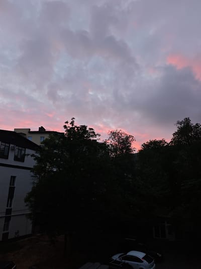The image captures a cityscape at dusk in Urozhainyi, Russia. The sky is filled with purple and pink clouds, suggesting a sunset. Silhouetted trees and the upper parts of buildings are visible against the sky. A white building with multiple windows is on the left, and below, a parking lot holds multiple vehicles. The overall scene is dark, with details obscured by shadows.