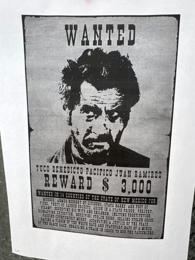 The image displays a black and white "WANTED" poster, likely a replica or prop, laid flat on a light-colored surface. The central subject is a stylized, grainy depiction of a man's face, presumably the wanted individual, Tuco Benedicto Pacifico Juan Ramirez. The poster is designed to mimic historical documents, with a distressed, aged appearance.
Visible text on the poster includes the large, bold word "WANTED" vertically on the left side. To the right, the name "TUCO BENEDICTO PACIFICO JUAN RAMIREZ" is prominently displayed, followed by "REWARD $ 3,000". Below this, it reads: "WANTED IN 14 COUNTIES OF THE STATE OF NEW MEXICO FOR MURDER, ARMED ROBBERY OF CITIZENS, STATE BANKS AND POST OFFICES, THEFT OF SACRED OBJECTS, ARSON IN A STATE PRISON, PERJURY, BIGAMY, DESERTING HIS WIFE AND CHILDREN, INCITING PROSTITUTION, KIDNAPPING, EXTORTION, RECEIVING STOLEN GOODS, SELLING STOLEN GOODS, PASSING COUNTERFEIT MONEY, ASSAULTING A JUSTICE OF THE PEACE, RAPING A VIRGIN OF THE WHITE RACE AND STATUTORY RAPE OF A MINOR OF THE BLACK RACE, DERAILING A TRAIN IN ORDER TO ROB THE PASSENGERS." The bottom right corner of the overall photo shows faint text "1 of 1". The setting depicted within the poster is the state of New Mexico. The photograph itself shows the poster on a flat, light surface, with parts of the surrounding white space visible.