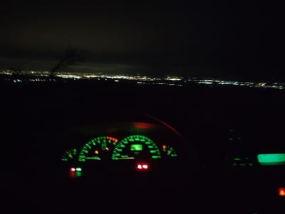 The image captures a nighttime view from the interior of a vehicle, overlooking the illuminated city of Balsha, Bulgaria, from an elevated vantage point. The foreground features the vehicle's dashboard, primarily displaying an instrument cluster with bright green backlighting for the speedometer and tachometer gauges. Several warning indicators are illuminated: a green light on the far left, an amber light centrally located within the cluster, and a red warning light positioned to its right. A faint green display and an additional red indicator are visible on the right side of the center console. The background reveals a panoramic expanse of the city lights of Balsha, appearing as numerous scattered points of yellow, white, and orange illumination against a dark, overcast night sky. A dark, bare tree branch is silhouetted against the sky in the upper-left portion of the frame.