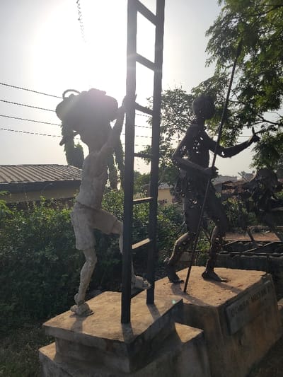 The image displays an outdoor public art installation in San Martín, Colombia, illuminated by bright daylight from the upper-left, causing lens flare. Two distinct, stylized human sculptures are positioned on individual, multi-tiered concrete pedestals. The sculpture on the left is a light-colored, possibly concrete, figure depicting a person in shorts carrying a dark, organic bundle on its head and left shoulder, with its right arm reaching towards a dark, vertical, ladder-like metal structure situated centrally between the figures. The sculpture on the right is a dark, open-form metal figure, depicting a person holding a long, thin spear or staff vertically with both hands. Both figures are presented in a walking posture. The pedestal supporting the right sculpture is inscribed with "OCHUBA NELSON C" and "2017". The background features dense green foliage and bushes, with a building exhibiting a corrugated metal roof partially visible in the far-left and a large green tree in the upper-right. Overhead utility lines are present in the upper-left quadrant of the frame.