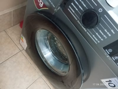 The image shows a close-up of a washing machine drum and control panel. The washing machine is gray and has a dark, reflective drum. The control panel has several options including "Стирка с паром" (Steam Wash), "Деликатная" (Delicate), "Синтетика" (Synthetics), "Очистка барабана" (Drum Clean), "Хлопок 20°C" (Cotton 20°C), "Быстрая 30'" (Quick 30'), "Быстрая 15'" (Quick 15'), "Эко 40-60" (Eco 40-60), and "Микс" (Mix). There is also a power button icon. A red object is partially visible on the floor to the left of the washing machine. The floor appears to be tiled. A sticker with a date "17.06.2026 01:40" and "10 YEAR WARRANTY INVERTER" is visible on the lower right side of the washing machine. The image is taken from a slightly high angle. The scene is indoors, likely in a home. The location is specified as Ohangaron, Uzbekistan.
