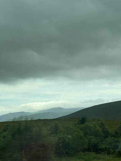 The image depicts a cloudy sky over a vast expanse of rolling green hills. In the foreground, dense foliage and trees blur due to motion, suggesting the photograph was taken from a moving vehicle. The middle ground features a textured green hillside, dotted with smaller shrubs and trees. Beyond this, layers of distant mountains fade into the hazy atmosphere, their slopes covered in muted green and patches of lighter color, possibly indicating sparse vegetation or snow. The sky is dominated by heavy, dark grey clouds, with lighter, diffused clouds and patches of pale blue peeking through in some areas. The overall impression is a moody, atmospheric landscape. The location is identified as Drimnaraw, Ireland. There are no people or specific actions depicted in the scene.