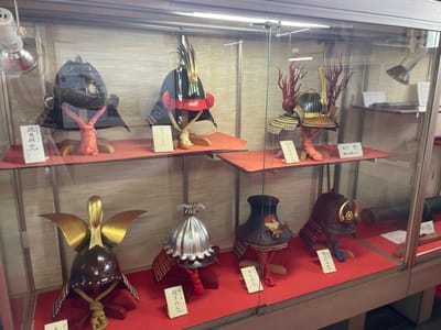 This image captures a display of samurai helmets within a glass-fronted cabinet. The cabinet is situated in what appears to be a museum or historical exhibit. The helmets are varied in design, with some featuring elaborate crests and decorations, while others are more minimalist. Each helmet is mounted on a stand, and many have small explanatory cards in front of them, written in Japanese.

The lighting in the cabinet is artificial, with a spotlight visible at the top right, casting illumination on the objects. The background within the cabinet is a textured, light-colored wall. The overall presentation suggests a curated exhibition of cultural artifacts.

Based on the presence of samurai artifacts, this display is likely located in a region with significant historical ties to the samurai class. Iga, Japan, is renowned for its association with ninjas and samurai, making it a plausible location for such an exhibition. While no people are visible in the image, the context of a museum suggests a public space.