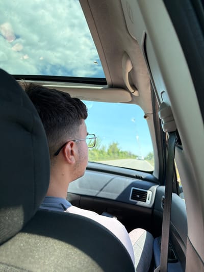 The image shows a person lying down in a car. The person is wearing glasses and has dark hair. The person's head is resting on a black car seat. A seatbelt is visible. The car's interior is visible, including the roof, handle, and air vent. Outside of the car is a blue sky, trees, and the edge of a road. There is no information to determine if the person is in Belovitsa, Bulgaria.