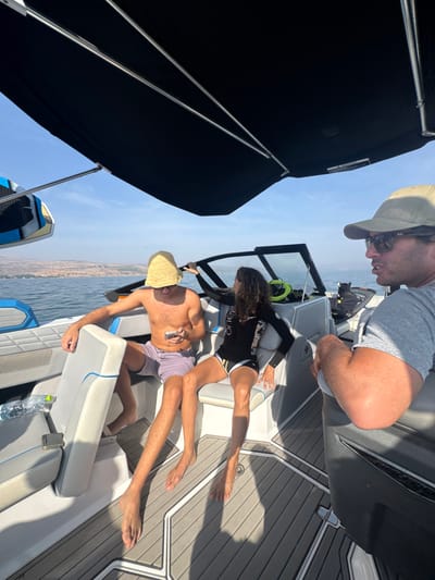 The image displays three individuals aboard a white and grey recreational motorboat on the Sea of Galilee, located near Migdal, Israel, under a clear blue daytime sky. The boat features a black canvas bimini top, white and grey marine-grade upholstered seating, and grey patterned non-skid flooring.
On the left, a shirtless male subject wears purple swim shorts and a straw hat pulled down over his face, obscuring it. He is seated on a bench, holding a silver smartphone and looking down at it. A clear plastic water bottle rests on the boat floor beside him.
In the center, a female subject with dark curly hair wears a black long-sleeved wetsuit top with an "O'neill" logo and patterned shorts. She is seated on an adjacent bench, facing slightly towards the bow, with her right hand gripping a dark rail near the windshield.
On the right, a third male subject is partially visible, wearing a tan baseball cap, dark sunglasses, and a grey short-sleeved t-shirt. He is seated in a darker grey seat, observing something outside the frame or speaking.
A blue and black wakeboard is mounted on a rack on the port side of the boat. The background shows calm blue water extending to a distant shoreline characterized by rolling hills and scattered urban development.