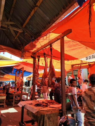 A bustling outdoor market scene is captured under a mix of corrugated metal and orange tarpaulin roofing. Pieces of raw meat hang from wooden supports, indicating a butcher stall. People are scattered throughout the scene, some shopping, others standing or walking. A man in a grey shirt leans against a post, observing the market activity. Tables with various food items are visible, suggesting a variety of vendors are present. The overall atmosphere is vibrant and lively, typical of a busy marketplace.