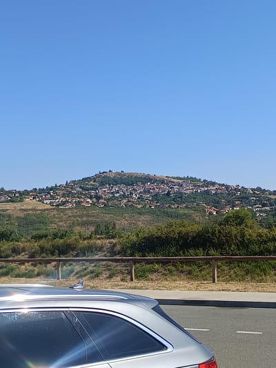 The image shows a panoramic view of the hilltop town of Monton, France, under a clear blue sky. The town, with its red-tiled roofs and white-walled buildings, is nestled on a verdant hillside dotted with trees. A prominent statue or monument is visible on the highest point of the hill. In the foreground, the roof and rear window of a silver car are visible, parked on the side of a paved road. A wooden fence runs horizontally across the middle ground, separating the road area from the hillside. The overall environment suggests a sunny day in a rural or semi-rural setting.