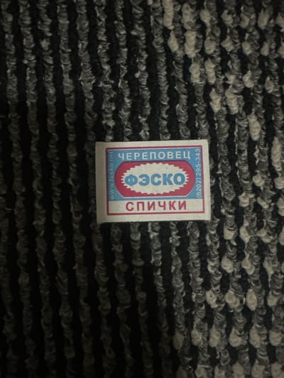 A close-up, top-down shot shows a matchbox with a label featuring text in Russian. The label is white with a red border and contains blue and red text and graphics. At the top, in blue, are the words "ЧЕРЕПОВЕЦ". Below that, inside an oval with a jagged red border, is the blue word "ФЭСКО". At the bottom, in red, are the words "СПИЧКИ". To the right of the oval, vertical text in parentheses reads "(8202) 295-343". To the left of the oval, vertical text reads "www.fasko.net". The matchbox is resting on a textured dark gray and white surface, possibly carpet or fabric, with a striped pattern. The image is cropped and does not provide any context about its location in Samarkand, Uzbekistan, or any people, actions, or interactions.