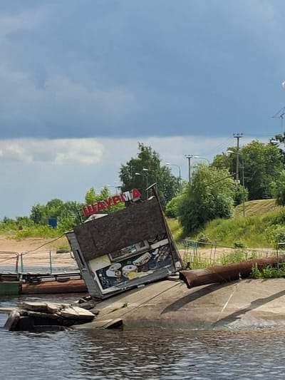 In the city of Verhnij Uslon, Russia, a dilapidated kiosk, seemingly a food stand with the Cyrillic word "ШАУРМА" (SHAURMA) prominently displayed in red lettering, is tilted precariously on a concrete embankment. The structure appears abandoned, with its wooden facade showing signs of wear and tear, and a graphic depiction of food items visible on its side. The kiosk is situated near the water's edge, with debris, including what appear to be concrete blocks and a rusty metal pipe, scattered around its base. The surrounding environment features lush green trees and vegetation, with a cloudy sky overhead. Electrical poles and wires are visible in the background, suggesting the presence of a developed area. The scene conveys a sense of neglect and disrepair.